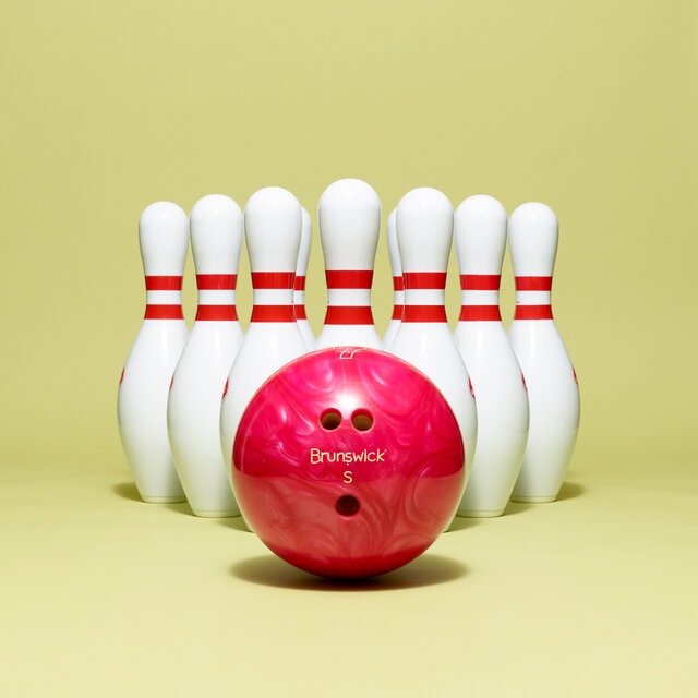 A red bowling ball sits in front of 10 bowling pins in a triangle formation on a yellow background. 