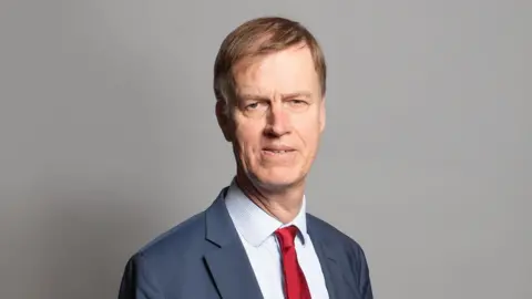 Department of Work and Pensions Minister for disability health and work, Sir Stephen Timms looks at the camera in front of a plain grey background. He is wearing a red tie to represent his position in the Labour party and is wearing a grey suit with a pin striped, white shirt. He has brown hair swept to one side.