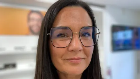 A woman with long brown hair and dark wire framed glasses is looking a the screen, smiling. You can see blurred out glasses in the background.