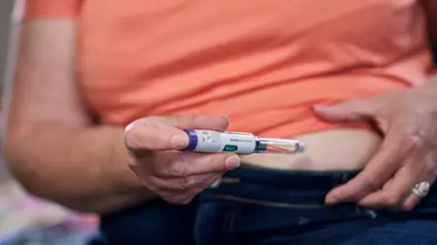 Getty Images A close-up of an overweight woman wearing jeans and an orange top, who is injecting a diabetes drug into her stomach using an injectable pen.