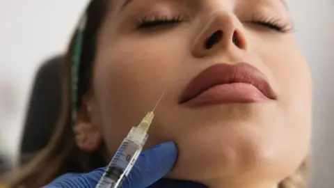 Getty Images A close-up of a woman's face as she lies back, waiting for a filler injection in her upper lip, as a needle hovers above her mouth