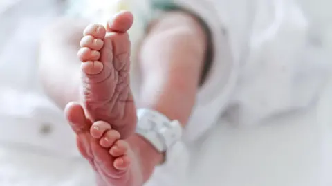 Getty Images A close-up shot of a baby's feet. The infant is lying down and has a white hospital tag around their ankle. The rest of the baby is blurry.
