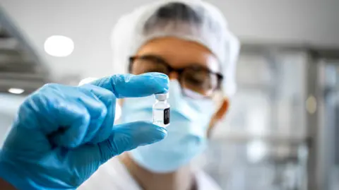 Getty Images A scientists wearing blue gloves, lab coat and hair net holds a vial containing a clear liquid