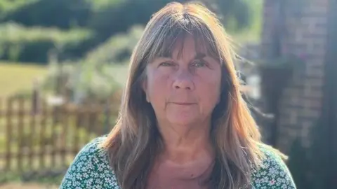 Eady family Claire Eady looks at the camera while wearing a green floral print top. She has shoulder length light brown hair and a fringe and is standing outside with blurred greenery visible in the background.
