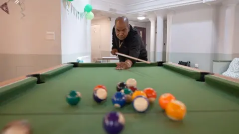 A man holds a pool cue after taking a shot at a pool table. There are balls in the foreground