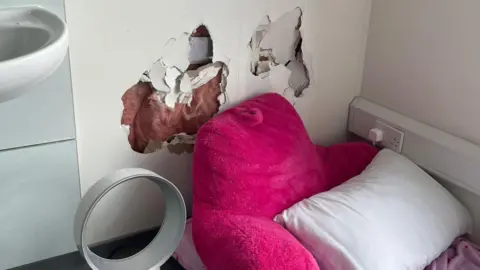 BBC A view of a wall with damage. In front is a pink bean bag, to the side is a fan and a sink.