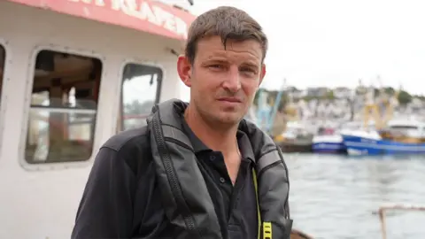 BBC Tom Parker is staring straight into the camera. He is standing on his fishing boat in the harbour, and wearing a black T-shirt and a black plastic life jacket. Behind him is Brixham harbour, with fishing boats in the harbour. 