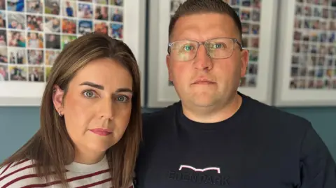 A woman and a man are standing in a room looking at the camera. Both have neutral expressions on their faces. To the left is the woman, who is called Sara Watson. She has long brown hair and is wearing a white top with red horizontal stripes. David Watson is on the right,. he is wearing glasses and has short, spiked hair. He is wearing a black t-shirt. 