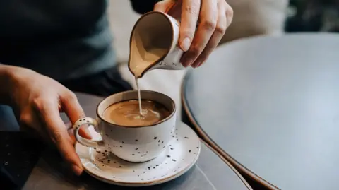 Getty Images Milk from white jug with brown speckles on it being poured into a cup with black coffee in it. Cup is on a saucer and matches the jug. We can see two hands holding cup and jug. 