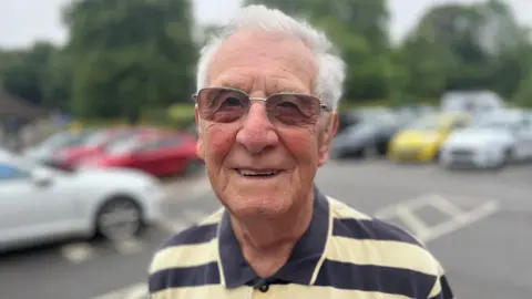Alvin Trevenna is wearing sunglasses and facing the camera while wearing a lemon and navy striped polo short, with blurred out cars in a car park visible behind him.