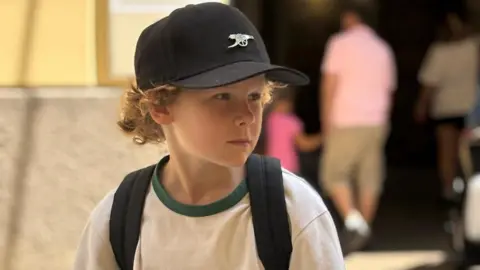 Handout A light haired boy wearing a white t-shirt and black cap looks into the distance.