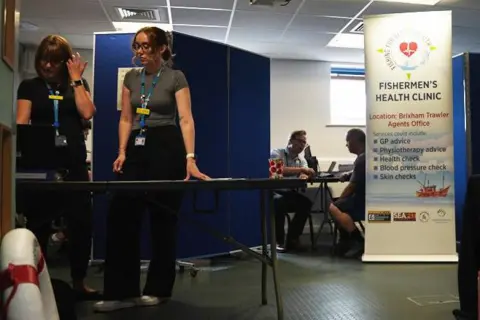 A wide shot showing the entrance to the fishermen's health clinic in Brixham trawler agent's office. There are two receptionists in front of a table in the room which is divided up by temporary blue screens. A doctor is in the middle cubical speaking to a patient. A temporary sign lists the various services on offer including GP access, blood pressure checks and skin cancer checks.  