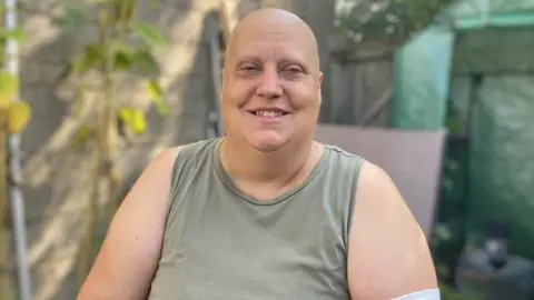 BBC Faye Woodley looking ahead and smiling. She is sat down in a sunny garden. She has no hair and is wearing a light green tank top.