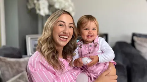 Baby Saskia is being held by her mother Scarlett Jones, who has long blonde hair and is wearing a candy pink shirt with white stripes. Baby Saskia is wearing pink and white striped dungarees and is holding an egg toy. They are both really smiling. 
