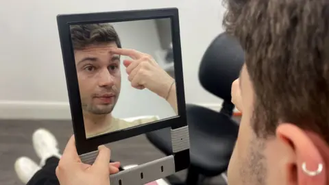 Ruth clegg/BBC Ven Grecu is looking in the mirror. He is a 24 year old man with brown hair and a faint beard. He is pointing at his forehead in the mirror.