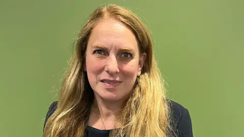 Leila Biant, with long, wavy blonde hair, stands against a plain green background. She is wearing a dark-colored top.