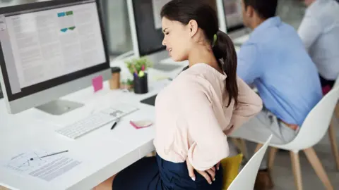 Getty Images Woman sitting at a chair holding her lower back and wincing in pain