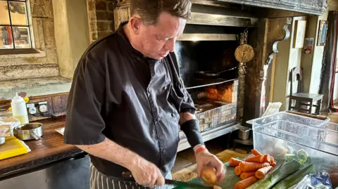NIKKI FOX/BBC Tony Henderson is in chef's clothing. A black chef's jacket and a striped pinny. He is standing in front of an open fireplace where they cook meat at the Gunton Arms near Cromer. He is standing in front of a green chopping board and is chopping onions. Tony often works in a customer facing position cooking meat. His left arm has an elasticated armband on it to protect his arm where the transfusion fistula goes into his arm.