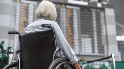 Getty Images Wheelchair user photographed from behind looks at a large departures board in an airport