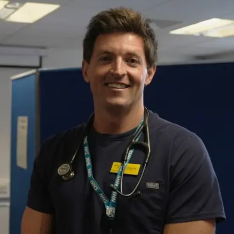 Dr James Gunning, an NHS GP, stands in the temporary fishermen's health clinic in Brixham. He is wearing an NHS T-shirt and has a stethoscope around his neck. Behind him is a receptionist looking at her computer and booking patients into the clinic who turn up for health checks.