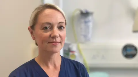 Dr Catherine Monaghan is standing by a hospital bed, out of focus behind her. She is wearing her dark blue medical scrubs uniform. Her blonde hair is tied back.