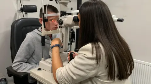 A boy is having a light shone into his eye as part of an eye examination. His head is resting on a metal frame and an Optometrist is examining his eye through a lens. The optometrist has long dark hair and a cream coloured jacket. Her back is to the camera. The boy has short dark hair and is wearing a grey coloured hoodie. 