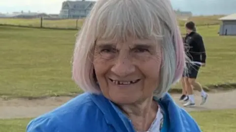 Claire Eady Geraldine Gibson smiles at the camera in a bright blue jacket. She has a short grey bob and fringe and grass and the sea is visible behind her.