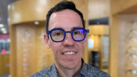 A man with short dark brown hair is smiling at the camera. He is wearing thick rimmed blue glasses and a paisley patterned shirt. 