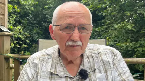 Stephen Evans is sitting outside in his garden, he is wearing a white and cream checked shirt with the top button undone and a microphone is affixed to his lapel. Behind him is what appears to be the end of a shed and some fencing and there are trees and bushes in the background.