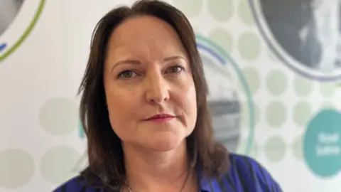Alison Hernandez, South West Police and Crime Commissioner, wearing a royal blue and black striped shirt looks directly at the camera with a neutral expression on her face.