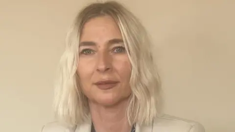 Tricia Roberts Tricia Roberts has shoulder-length blonde hair and is stood against a cream background. She is wearing a cream blazer and has a neutral expression on her face as she looks at the camera. It is a head and shoulders shot of her. 