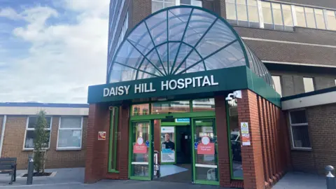 A large brown bricked building has a green banner on it's front that reads 'DAISY HILL HOSPITAL.' The banner sits at the entrance to the building below a glass archway which is encased in a green skeletal structure. They sit on-top of a red brick extension out the front of the brown bricked building. There is a black bench and a small green tree to the left of the entrance