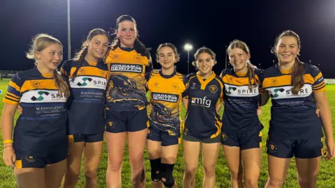 Seven members of the Worcester RFC Under 16 girls team are smiling as they pose for the photo. They are wearing blue and gold shirts and navy shorts. We have permission forms for each player. 