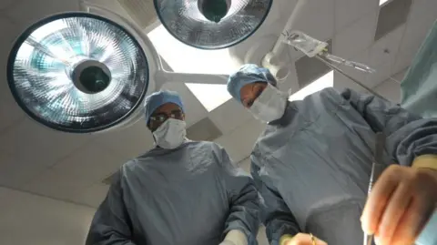 BBC Two surgeons in surgical gear and masks look on in an operating theatre with one of them using a scalpel