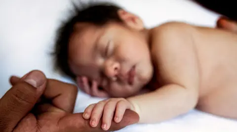 Getty Images New born baby asleep with hand outstretched and fingers curled round the finger of an adult. Baby has lots of dark hair and is resting their head on their other hand. 