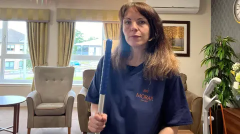 BBC Photo of Catriona in the care home. The photo is a selfie which appears to have been taken with the camera balanced n a table. The background shows the interior of the home with her cleaner's trolley behind. Catriona is dressed in a blue overall.