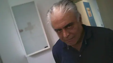 A man with grey hair stands in a clinic corridor, glancing towards a hidden camera while being covertly filmed describing illegal Botox practices.