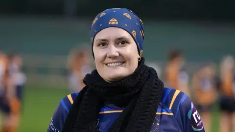 Jenni Hussain is wearing a hijab made out of navy material with a yellow Worcester RFC badge, a Worcester RFC shirt and a black wool scarf. She is smiling and standing on a rugby pitch