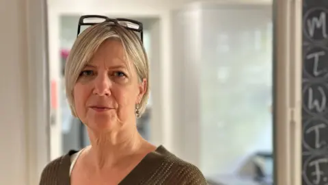 Sarah Milner looks straight into the camera. She has straight blonde hair in a smart bob and has her glasses pushed onto the top of her head. She is wearing a brown V neck top and earrings. She is standing in a doorway, with a blurred white wall behind her. 