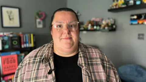 The picture is of a white woman with dark hair pulled back off her face, wearing clear frame glasses and a checked shirt with a black t-shirt underneath. She's stood in a room with books in the background and pictures on the wall.