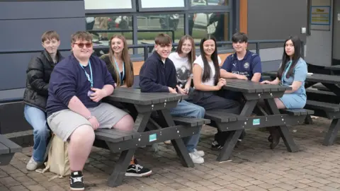BBC A group of male and female students, all aged 17, sit together, smiling, at two picnic tables in front of their college building