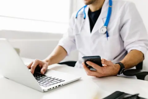 Getty Images Doctor in a white coat with a stethescope round his neck looking at a laptop with a phone in his left hand. You can only see the body and not the head