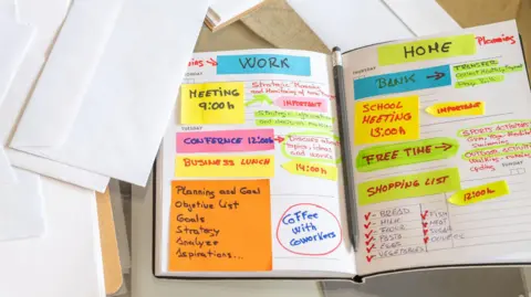 Getty Images Person is organizing their work and personal agenda in a notebook at home, using sticky notes and colorful markers to plan their week, surrounded by envelopes, a smartphone, and a notepad 