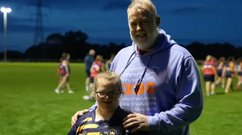 Elsie-May and her Dad David are standing on a rugby pitch. She has black rimmed glasses and wears a blue top with yellow stripes. He wears a light blue hoodie with orange/white writing and has short white hair and a beard.