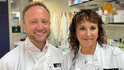 BBC/Fergus Walsh Two scientists, Professors Ed Wild and Sarah Tabrizi, are pictured from the shoulders up. They are both smiling and wearing white lab coats. Prof Wild on the left has a tightly cropped beard. Prof Tabrizi on the right has long wavy hair. They are clearly in a laboratory with scientific equipment on shelves in the background.