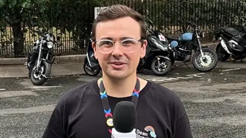 Josh Elgin/BBC Lawrence O'Connell is seen wearing glasses and a black T-shirt is holding a microphone while standing on a street, with several parked motorbikes and a fence with greenery in the background.