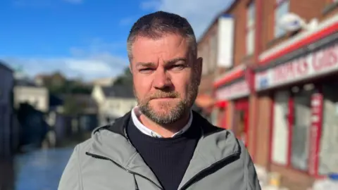 PA Media A grey haired man wears a grey waterproof jacket, black jumper and white shirt. He has a short brown beard. Over his right shoulder is an out-of-focus street with grey buildings. Over his left is a row of bricked properties, the one closest to him has a red shop front and glass windows. 