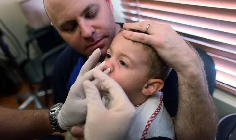 Getty Images Young boy wearing a grey t-shirt and white bib sits on man's lap to receive the flu nasal spray.