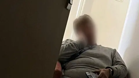 Supplied A person wearing a grey jumper and blue lanyard sits in a chair. They have their head tilted to one side and resting on the palm of their hand, seemingly asleep. 