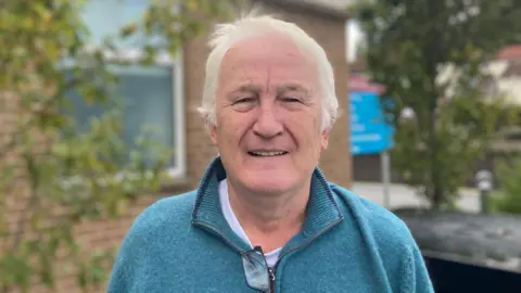 A white haired man wears a teal quarter zip jumper and a white t-shirt. His black lensed clear spectacles hang from the half zipped zip on his jumper. Behind him is an out-of-focus red bricked building with a translucent window over his right shoulder. There are also two small green tree's either side of him.